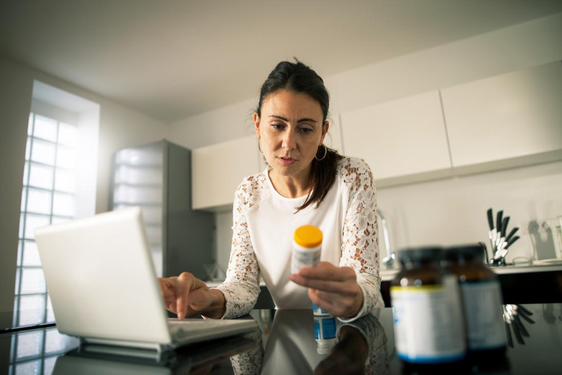 Woman in kitchen on laptop holding prescription pill bottle and reading label