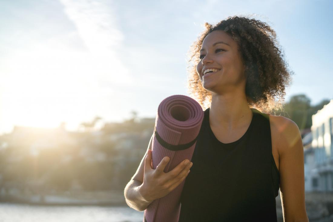 Woman smiling outdoors with yoga mat in sunshine.