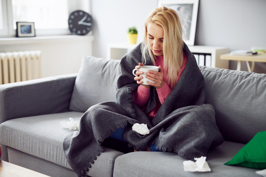 Woman with cold or flu sitting under blanket with mug of tea and tissues
