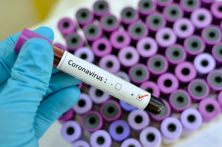 A gloved hand holding a blood sample vial marked Coronavirus positive